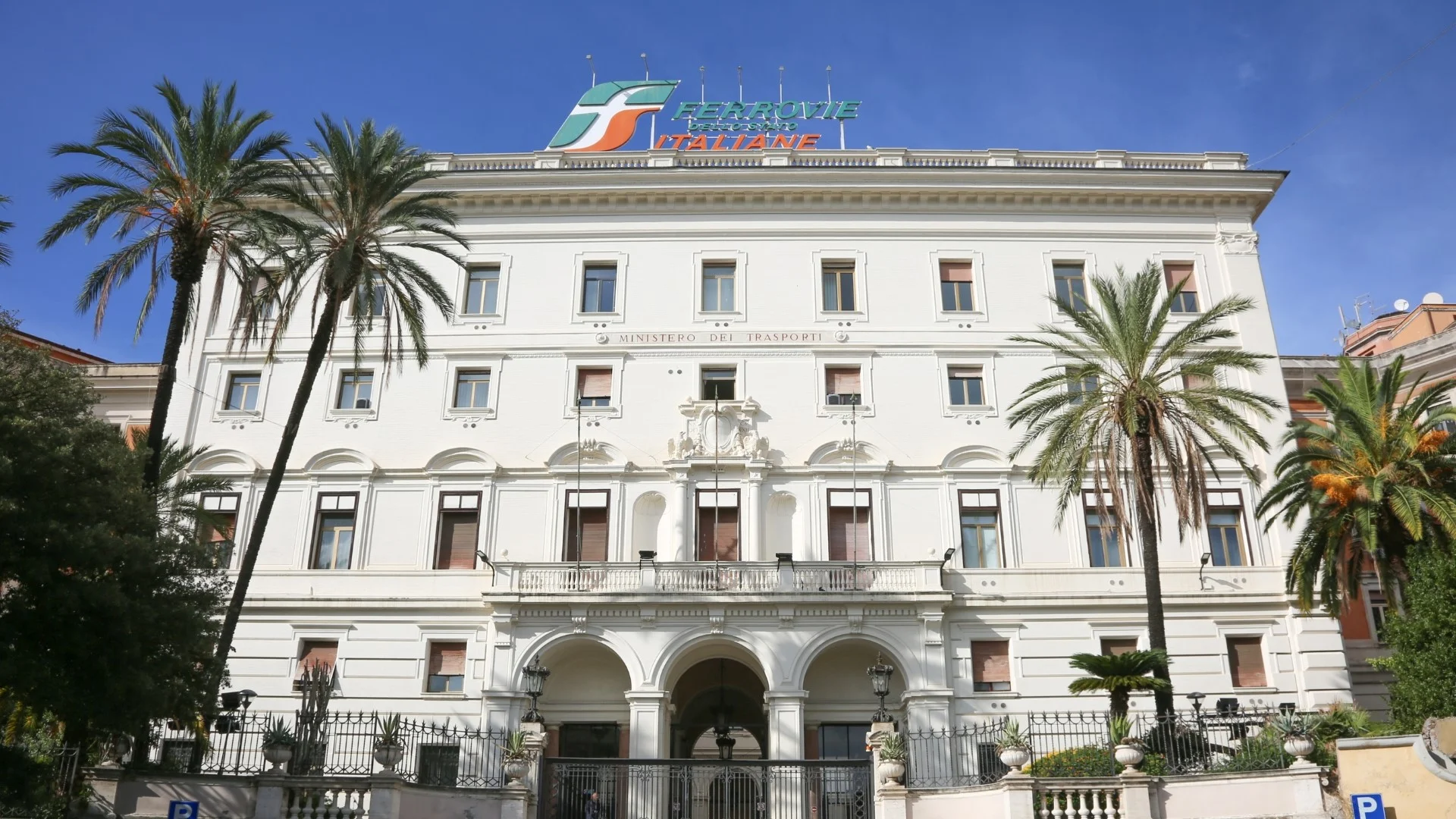 Historisches Verwaltungsgebäude mit Palmen vor dem Eingang, Fassade in klassischem Stil und großem Logo von Ferrovie dello Stato Italiane auf dem Dach.